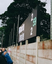A large digital scoreboard at an outdoor sports venue displays a score of 2-2. Spectators are gathered, observing the event with interest. The scene is enclosed by a tall metal fence and surrounded by lush green trees, indicating a natural setting. Overcast skies add a muted tone to the environment.