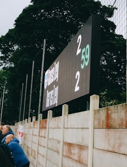A large digital scoreboard at an outdoor sports venue displays a score of 2-2. Spectators are gathered, observing the event with interest. The scene is enclosed by a tall metal fence and surrounded by lush green trees, indicating a natural setting. Overcast skies add a muted tone to the environment.