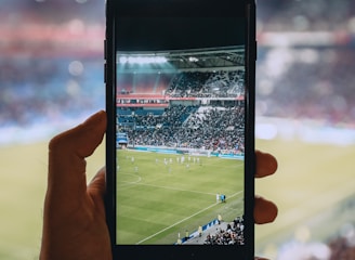 A hand holding a smartphone captures a live view of a soccer stadium filled with fans and players on the field. The image on the smartphone screen is crisp, showing a wide-angle view of the game in action.