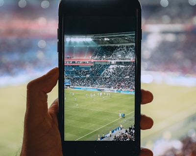 A hand holding a smartphone captures a live view of a soccer stadium filled with fans and players on the field. The image on the smartphone screen is crisp, showing a wide-angle view of the game in action.