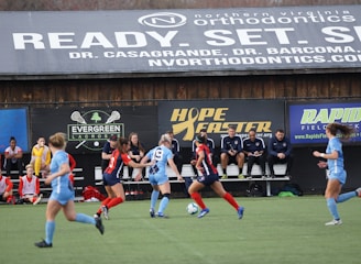 Players engage in a competitive soccer game on a field while others are seated on the bench watching attentively. The action is taking place in front of a wooden structure displaying multiple advertisements, including orthodontics, lacrosse, and field hockey.