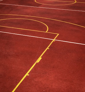 A sports court with a reddish-brown surface and bright yellow and white lines. The lines form geometric shapes, including rectangles and circles, suggesting markings for games like basketball or netball.