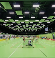 A sports hall with bright green flooring and ceiling panels. There is a game in progress with players moving quickly, suggesting a fast-paced sport like floorball or indoor hockey. Advertising banners line the walls, and a scoreboard is visible in the background.