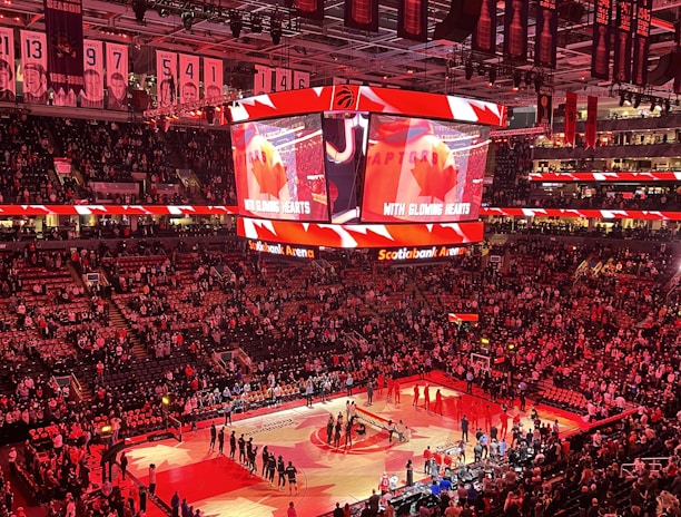 An indoor sports arena filled with spectators and a basketball court illuminated by red lighting. The scoreboards and video screens display promotional content, with numerous banners and championship flags hanging from the ceiling. Players and officials are gathered on the court, possibly in preparation for a game or event.