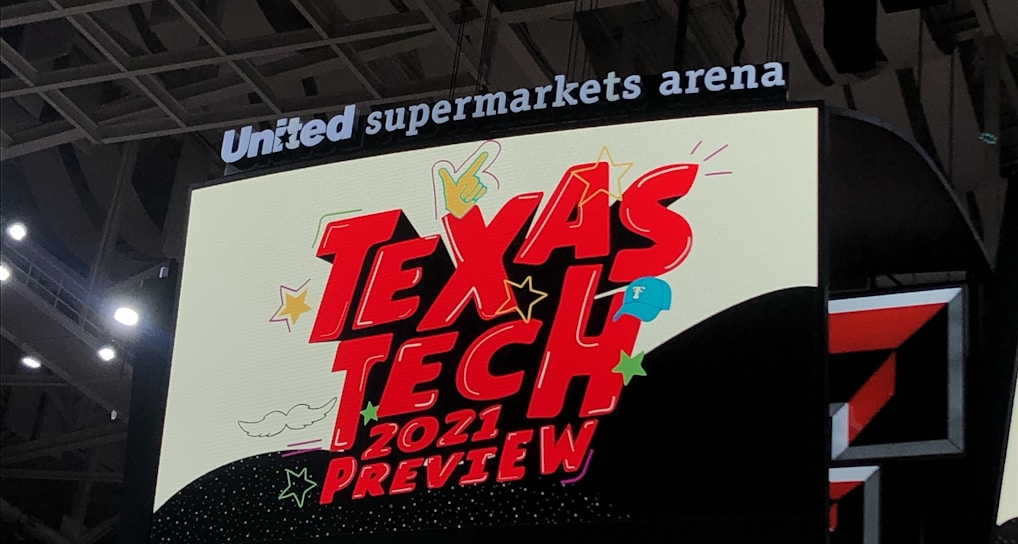 A scoreboard displaying the text 'Texas Tech 2021 Preview' in bold red letters with decorative elements like stars and clouds. The background is predominantly dark with bright accent colors, and the setting is within an indoor sports arena, specifically the United Supermarkets Arena.