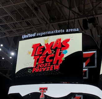 A scoreboard displaying the text 'Texas Tech 2021 Preview' in bold red letters with decorative elements like stars and clouds. The background is predominantly dark with bright accent colors, and the setting is within an indoor sports arena, specifically the United Supermarkets Arena.