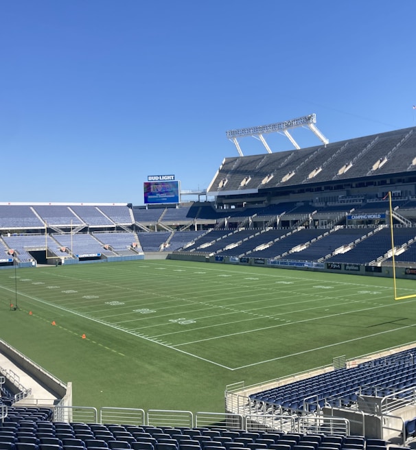 A large, empty sports stadium with blue seats surrounding a well-maintained green grass field. Bright sunlight creates clear shadows, and there are two goal posts on opposite ends of the field. A big electronic scoreboard displays some graphics, and the sky is clear and blue.