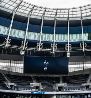 A modern stadium interior featuring a large digital screen with a logo prominently displayed. The seating areas are empty, with dark-colored seats and some structural elements like beams and glass walls visible. A combination of dark blue and neutral tones are prominent.